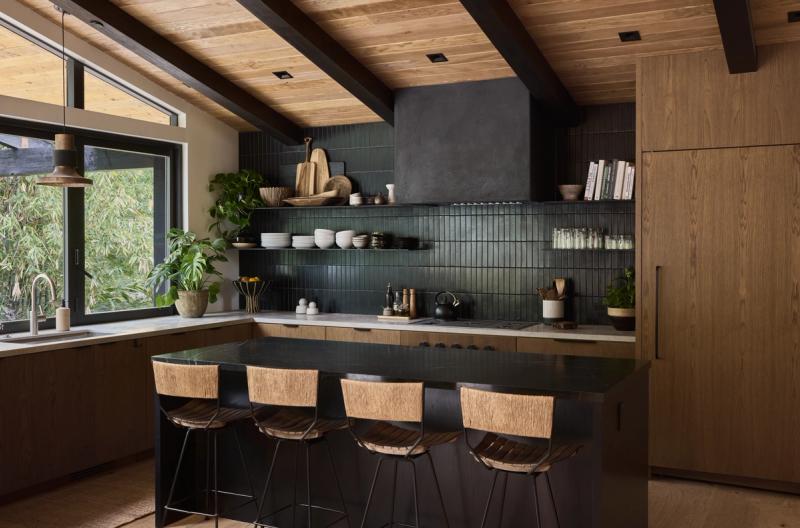 A modern kitchen featuring warm oak cabinetry, dark green vertical tile backsplash, open floating shelves, a black stone island with woven bar stools, and exposed wood beam ceiling.