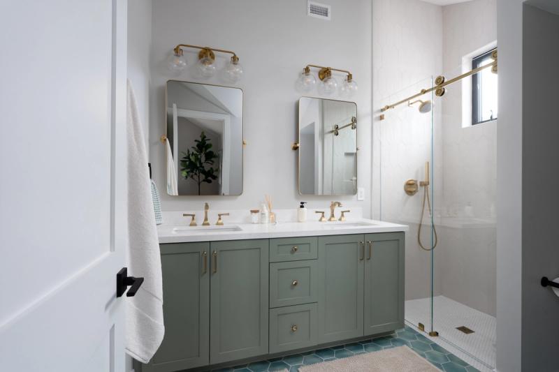 A bright, modern bathroom featuring a sage green double vanity with white marble countertop, gold brass fixtures and mirrors, hexagonal teal floor tile, and a frameless glass walk-in shower with brass hardware.