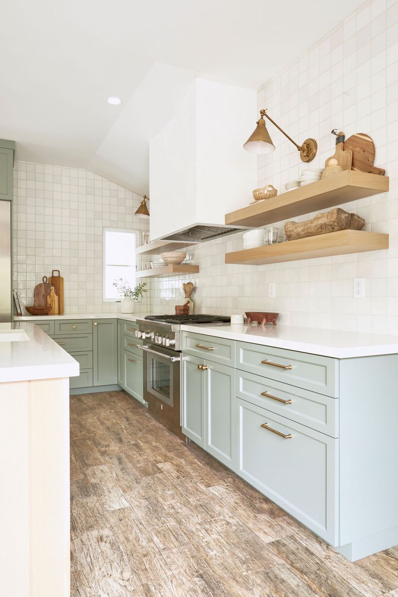 A bright, airy kitchen featuring sage green shaker-style lower cabinets with brass pulls, white quartz countertops, open oak floating shelves, white square tile backsplash, and wood-look tile flooring.