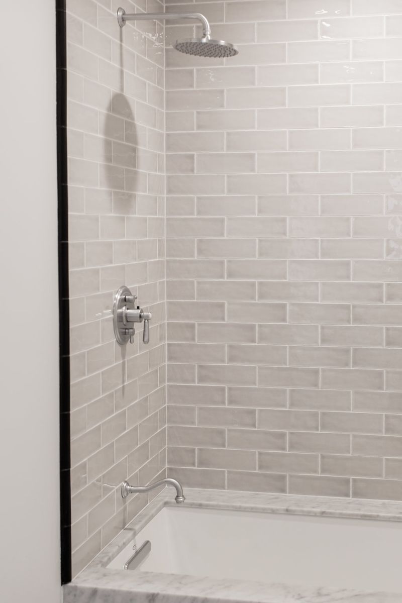 A clean, elegant shower and bathtub enclosure featuring light gray marble subway tile walls, a round rain showerhead, polished chrome fixtures, and a marble-edged soaking tub.