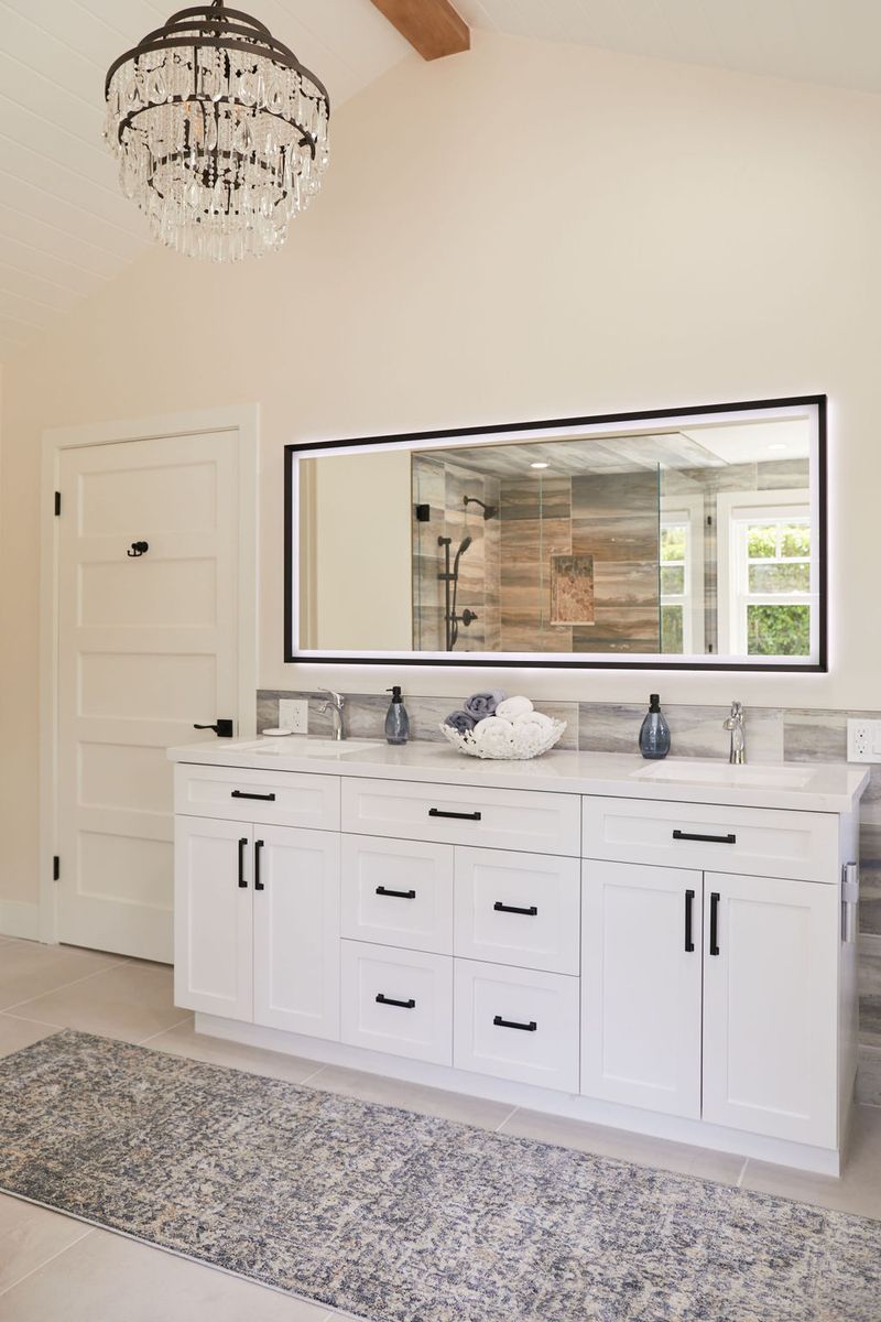 A bright, spacious master bathroom featuring a large white shaker-style double vanity with black hardware, a wide LED-backlit mirror, marble countertops, and an elegant crystal chandelier above a patterned area rug.