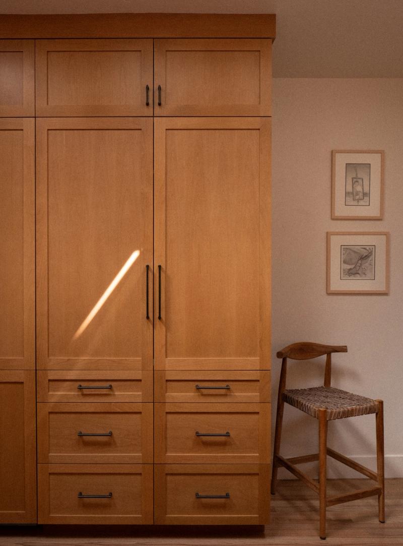 Tall custom cabinetry in warm honey-toned wood with panel-ready refrigerator doors, upper cabinet storage, and multiple lower drawers with black bar pulls, beside a woven-seat wood stool and framed artwork.