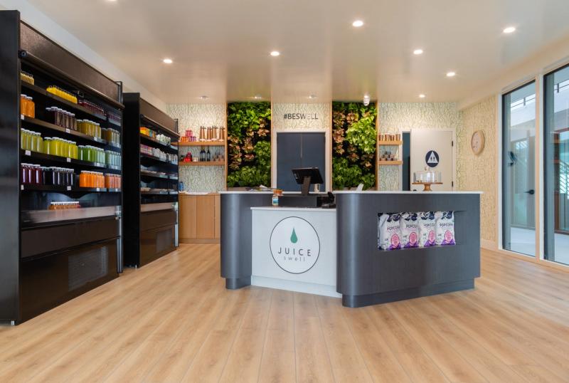 A modern juice bar interior featuring a curved gray service counter with the Juice Swell logo, floor-to-ceiling product shelving with colorful bottled juices, and a living green plant wall with the hashtag #BESWELL displayed above.