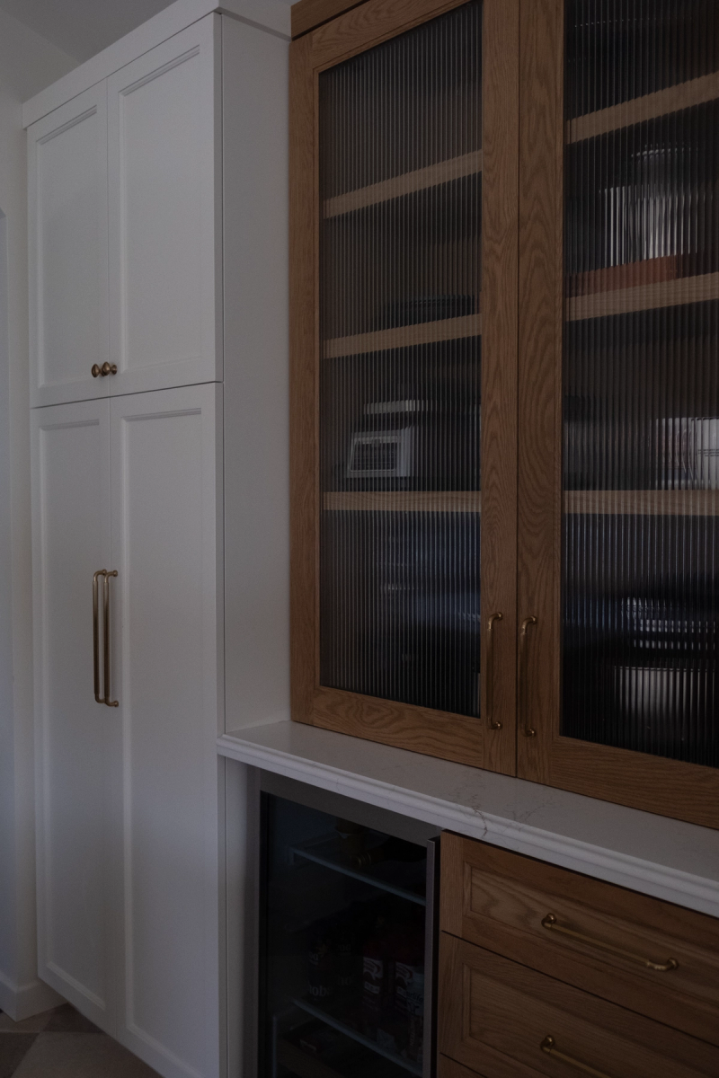 A butler's pantry featuring warm oak cabinets with fluted glass doors and brass hardware alongside tall white painted cabinetry, with a built-in wine cooler and marble countertop.