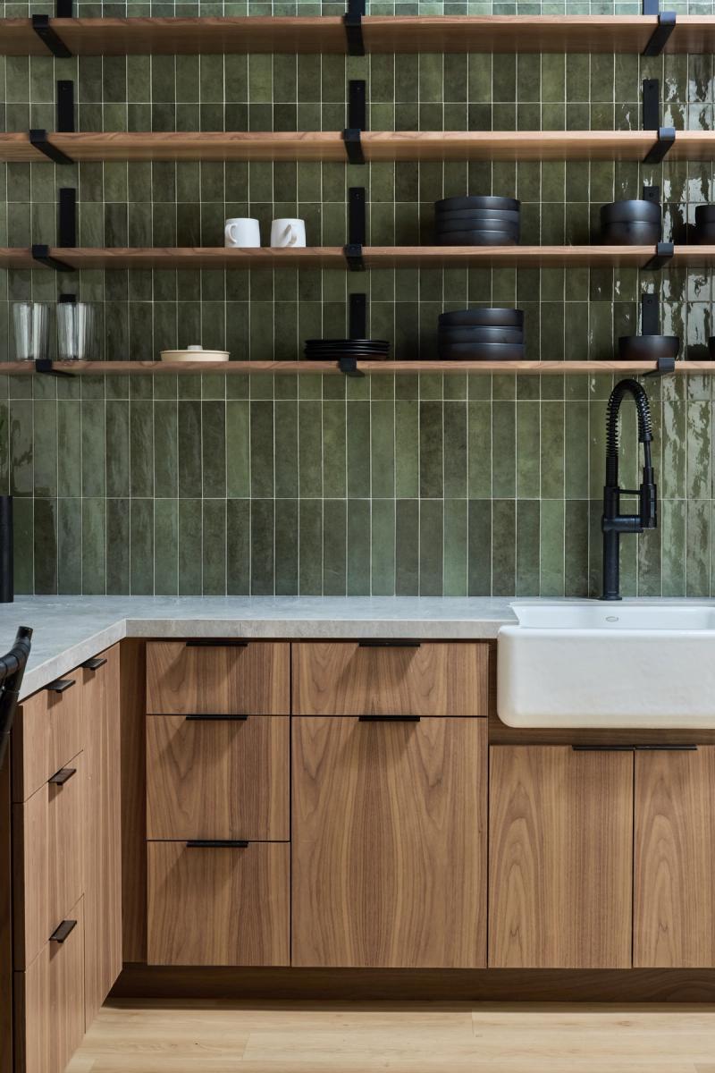 A modern kitchen featuring warm walnut cabinets with matte black hardware, a white farmhouse sink, a green vertical subway tile backsplash extending floor to ceiling, and open wood shelving displaying black dishware and white mugs.
