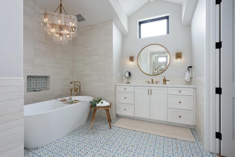 A bright, elegant master bathroom featuring a white shaker-style vanity with brass hardware, a freestanding soaking tub, patterned mosaic floor tile, and a crystal chandelier. The space includes a round brass-framed mirror, wall sconces, and decorative tile niche accents.