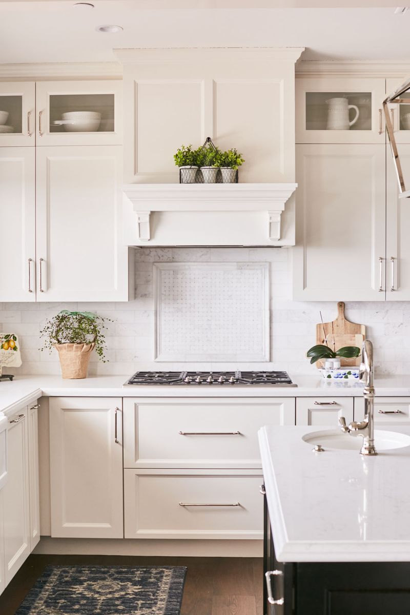 A bright, all-white traditional kitchen featuring custom inset cabinetry with polished nickel hardware, an elegant mantel-style range hood, marble backsplash, and white quartz countertops. Glass-front upper cabinets display white dishware and the space is accented with potted greenery.