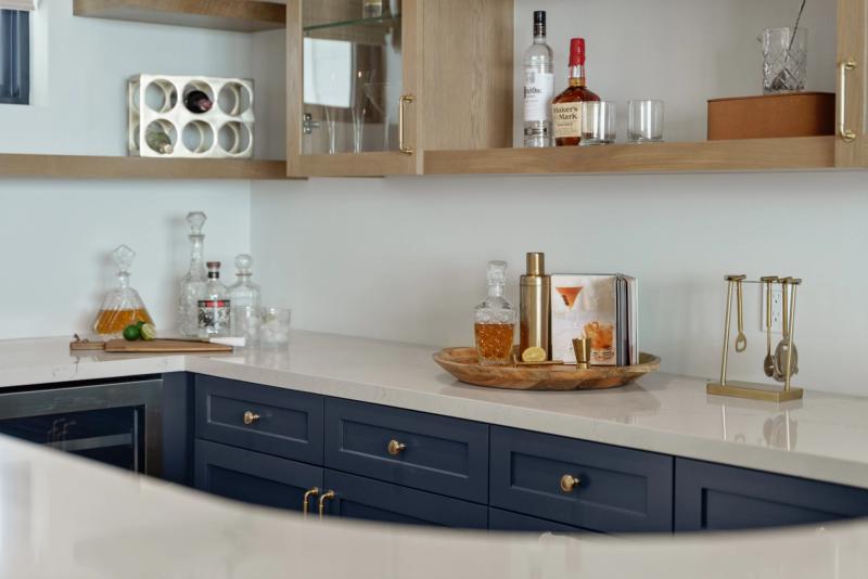 A stylish home bar area featuring navy blue shaker-style lower cabinets with brass hardware, white quartz countertops, and open natural oak upper shelving stocked with spirits, decanters, and glassware.
