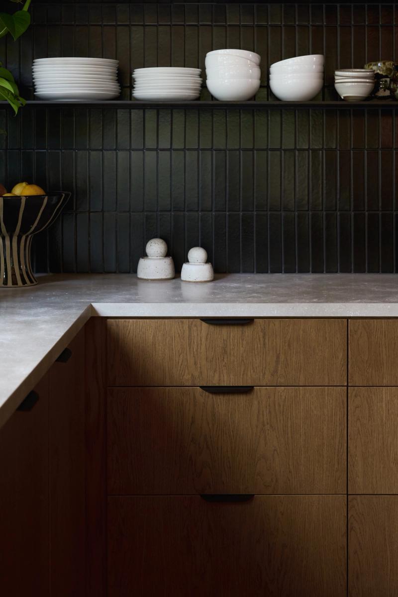A modern kitchen featuring warm oak drawer cabinets with black hardware, a light stone countertop, dark vertical tile backsplash, and open shelving displaying white dishes and bowls.