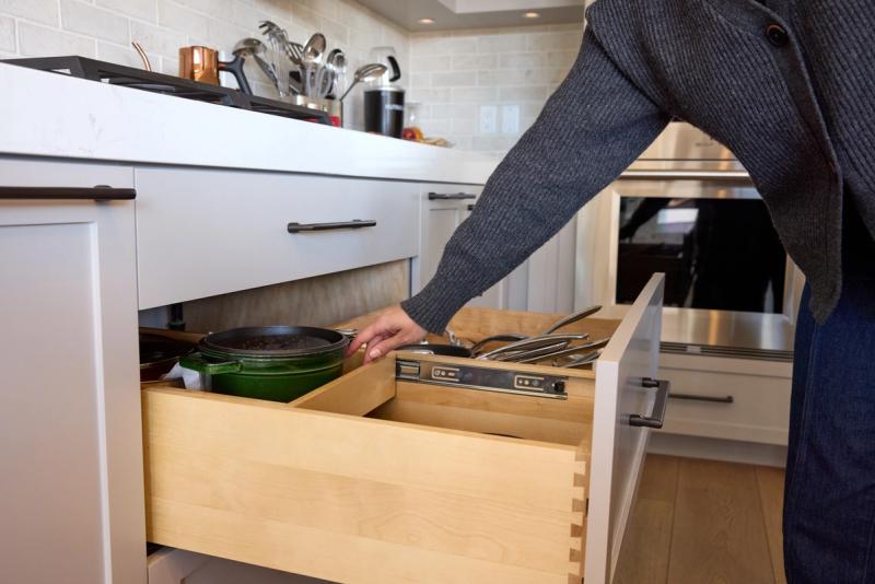 A person reaches into a large, deep pull-out base cabinet drawer storing cast iron and stainless cookware, with a secondary divided drawer insert above holding utensils and pan lids, set in a modern white kitchen.