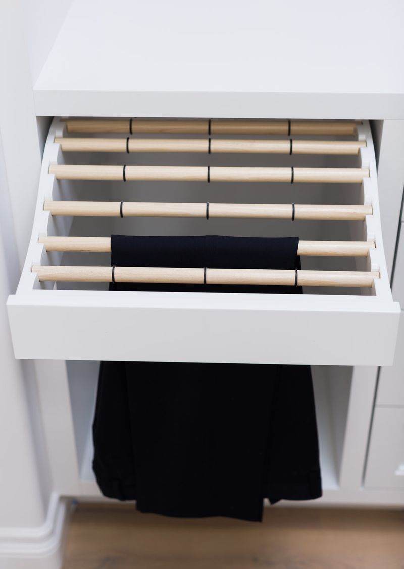 A white custom closet cabinet with an open pull-out drawer featuring horizontal wooden dowel rods for hanging and organizing pants, with a pair of black trousers draped over one of the rods.