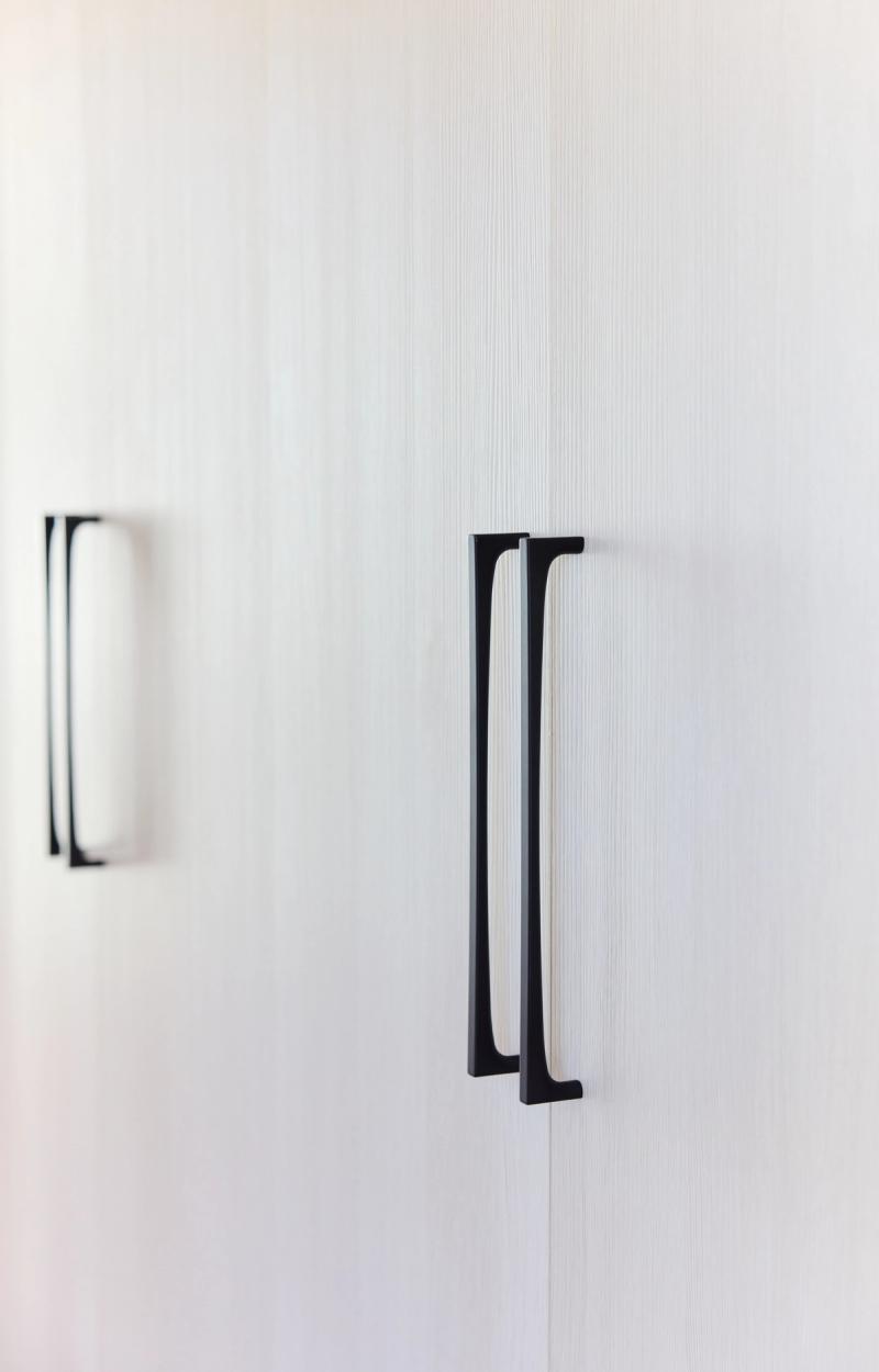 A close-up detail shot of sleek matte black elongated cabinet pulls mounted on white wood-grain cabinet doors, with a second handle visible in soft focus in the background.
