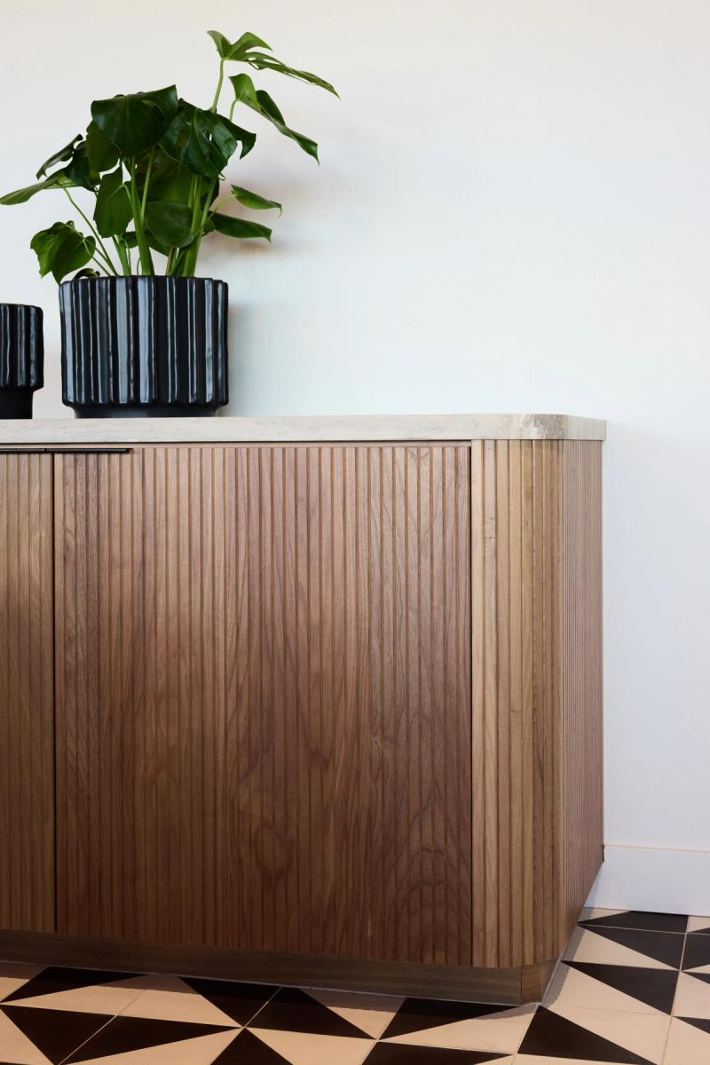 A close-up of a custom walnut sideboard featuring vertically fluted cabinet doors and a light stone top, styled with a black ribbed planter holding a green tropical plant, set on a geometric black and white tile floor.