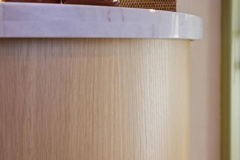 A close-up detail shot of a curved cylindrical cabinet base with light oak wood veneer and a white marble top surface.