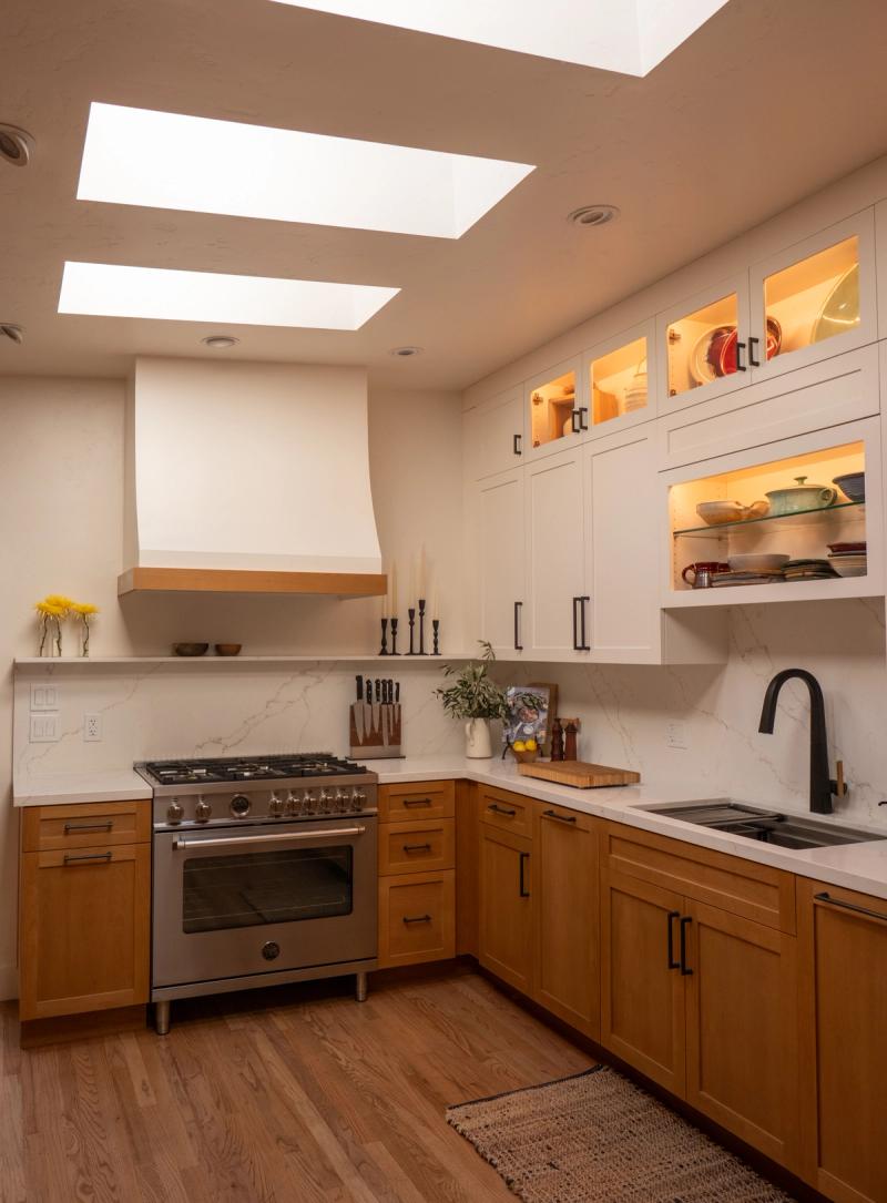 A warm, transitional kitchen featuring honey-toned lower cabinets, white upper cabinets with glass doors and interior lighting, a stainless steel range beneath a plaster hood, marble-look quartz countertops, and three skylights overhead.