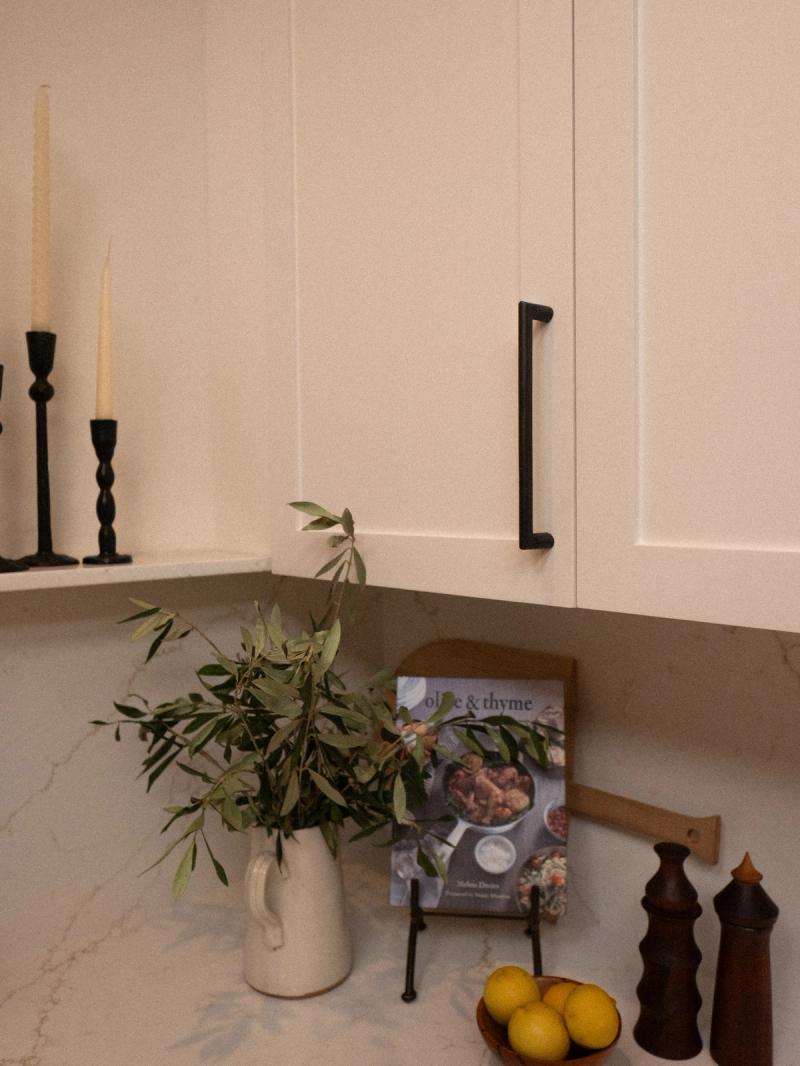 A styled kitchen corner featuring cream shaker cabinets with matte black pulls, a white marble countertop, and decorative accessories including an olive branch arrangement, lemons, wooden salt and pepper mills, and an Olive & Thyme cookbook.