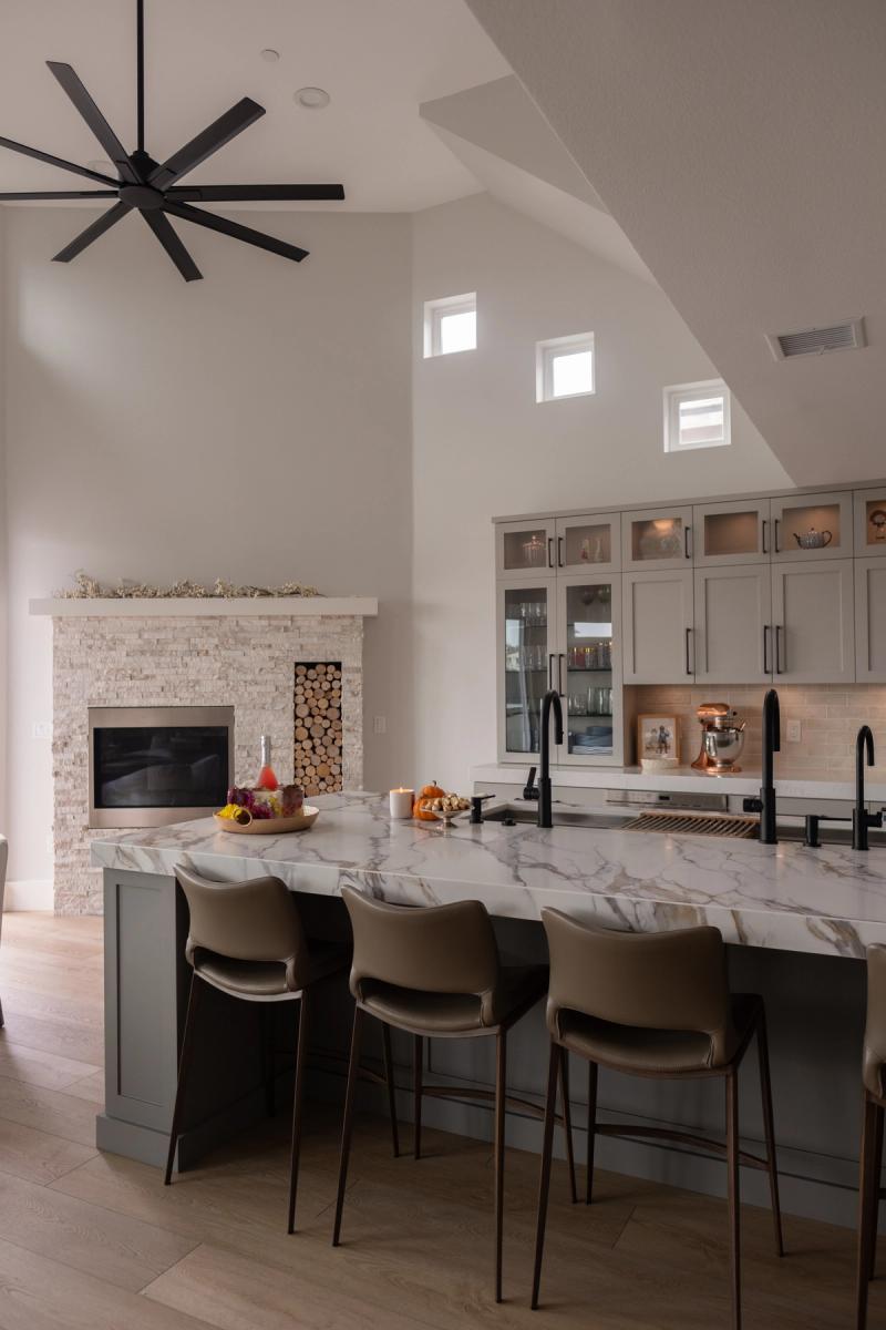 A spacious open-concept kitchen featuring a large marble-topped island with sage green base, gray shaker cabinets, black fixtures, and a stacked stone fireplace with firewood storage, topped by a dramatic vaulted ceiling with a large black ceiling fan.