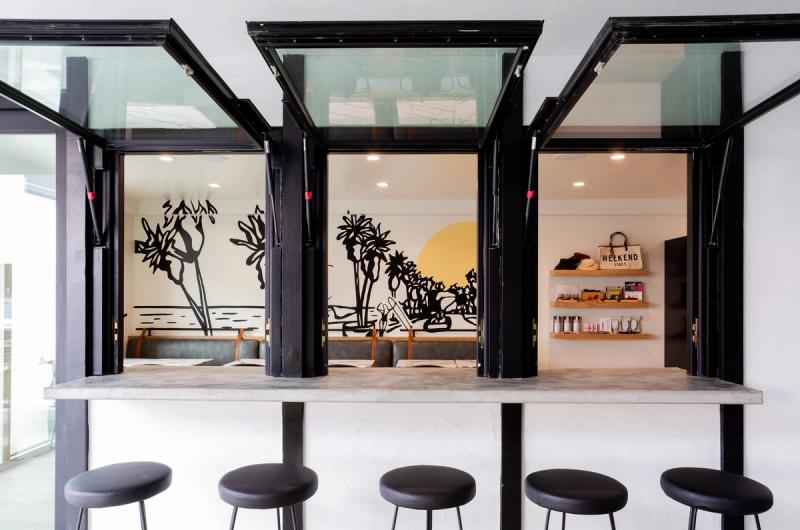 A concrete pass-through bar counter with black folding glass windows opens into a bright interior space featuring a bold black-and-yellow surf-themed mural and retail shelving with products and a tote bag.