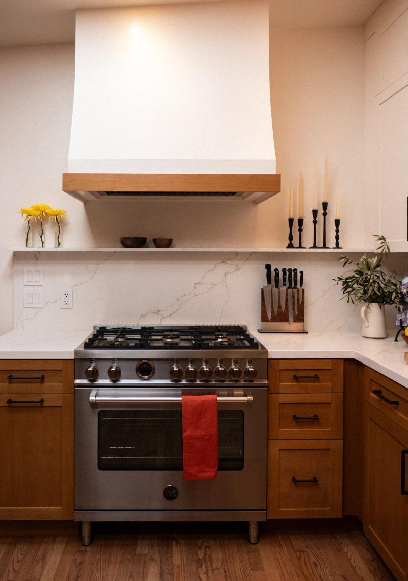 A custom kitchen featuring warm oak shaker cabinets with black hardware, a stainless steel professional gas range, white quartz countertops with marble-look backsplash, and a custom white and wood range hood.