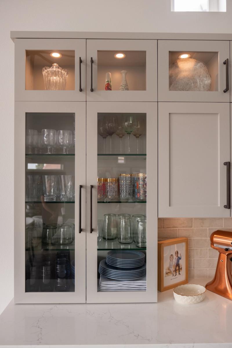 Tall gray shaker-style kitchen cabinets with glass doors display neatly organized glassware, wine glasses, and blue plates. Interior cabinet lighting illuminates decorative vases in the upper cabinets, with a copper KitchenAid mixer visible on the white quartz countertop below.