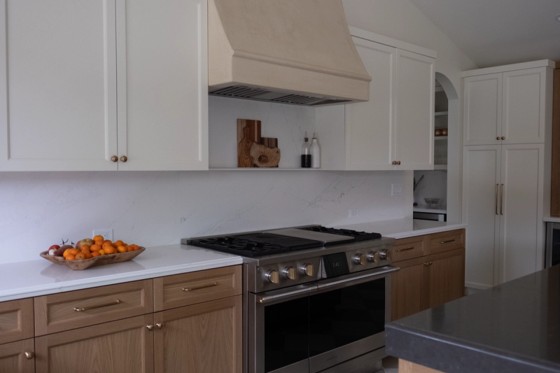 A bright, elegant kitchen featuring white upper cabinets and warm wood lower cabinets with brass hardware, a stainless steel gas range, marble-look quartz backsplash and countertops, and a custom plaster range hood.