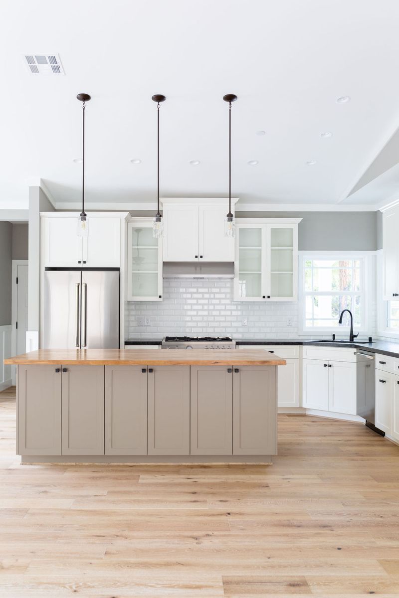 A bright, open kitchen featuring white shaker upper cabinets, a greige island with butcher block countertop, stainless steel refrigerator, white subway tile backsplash, black hardware, and light hardwood floors. Three pendant lights hang above the island.
