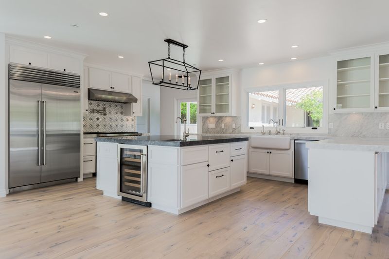 A bright, spacious custom kitchen featuring white shaker cabinets, a large center island with dark granite countertop and built-in wine cooler, farmhouse sink, patterned tile backsplash, and stainless steel appliances on light hardwood floors.