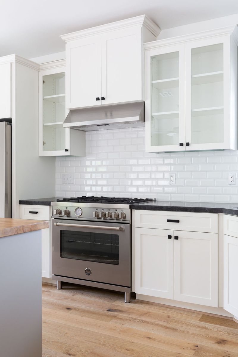 A bright white shaker-style kitchen featuring custom cabinetry with glass-front upper cabinets, black hardware, dark stone countertops, white subway tile backsplash, and a stainless steel professional range with hood.