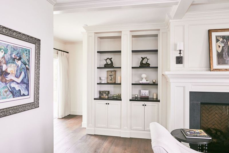 A bright living room featuring a white built-in bookcase with dark shelves decorated with figurines and candles, flanking a white fireplace surround with herringbone brick firebox.