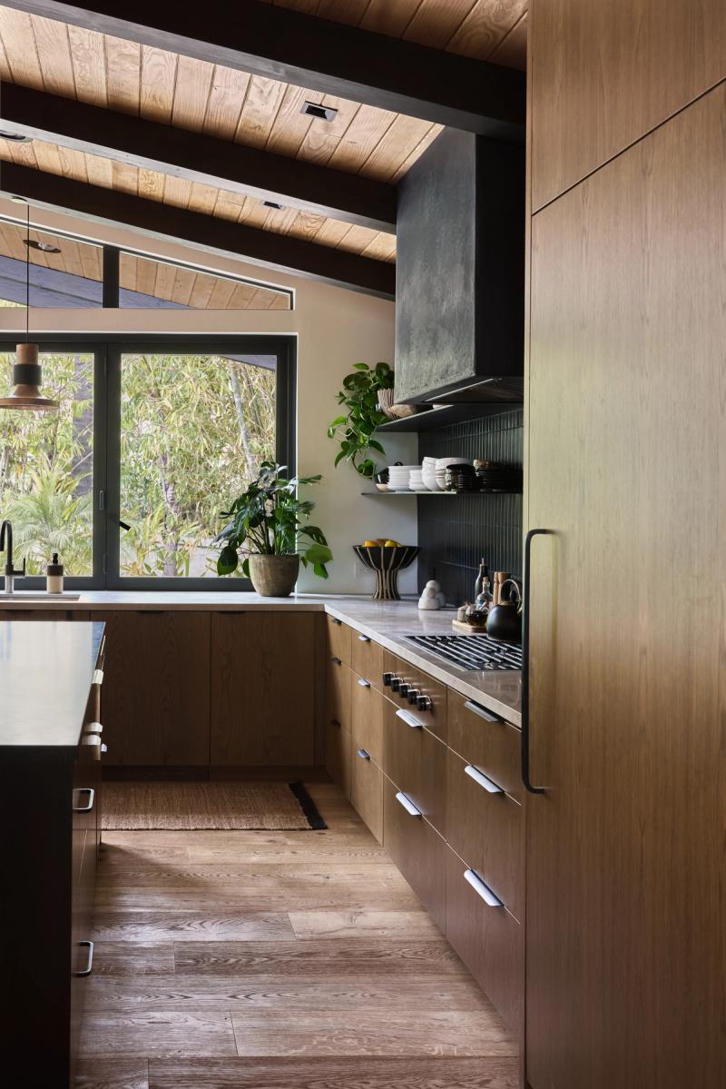 A warm mid-century modern kitchen featuring flat-front oak cabinetry, a concrete range hood, dark vertical tile backsplash, stone countertops, and exposed wood beam ceiling with large windows overlooking greenery.