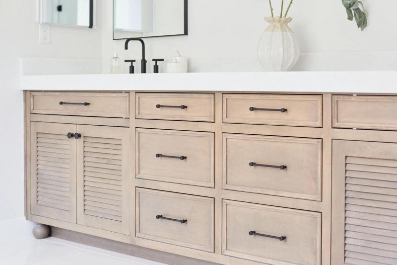 A wide custom bathroom vanity in a light driftwood finish featuring louvered cabinet doors, multiple drawers with matte black bar pulls, a white quartz countertop, and a black gooseneck faucet. A decorative white vase with greenery sits on the counter.