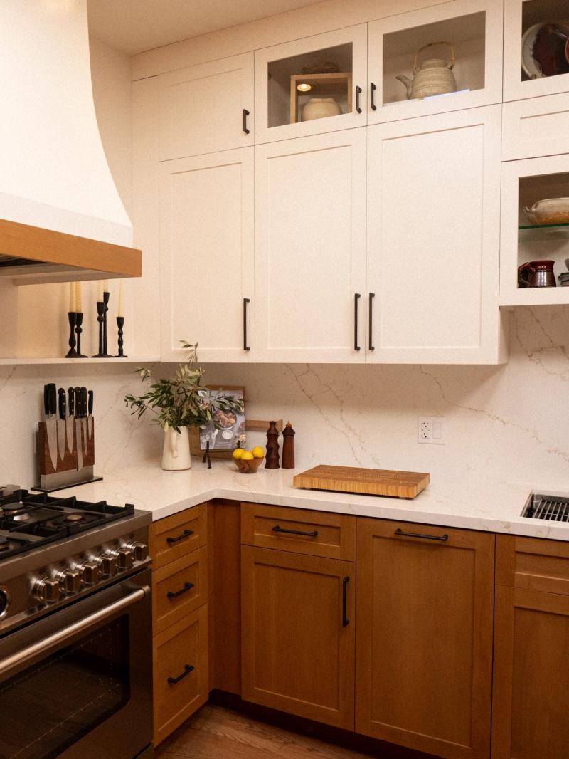 A custom kitchen featuring white shaker upper cabinets with glass-front doors and warm oak lower cabinets, black hardware, a marble-look quartz backsplash and countertops, and a professional stainless steel range.