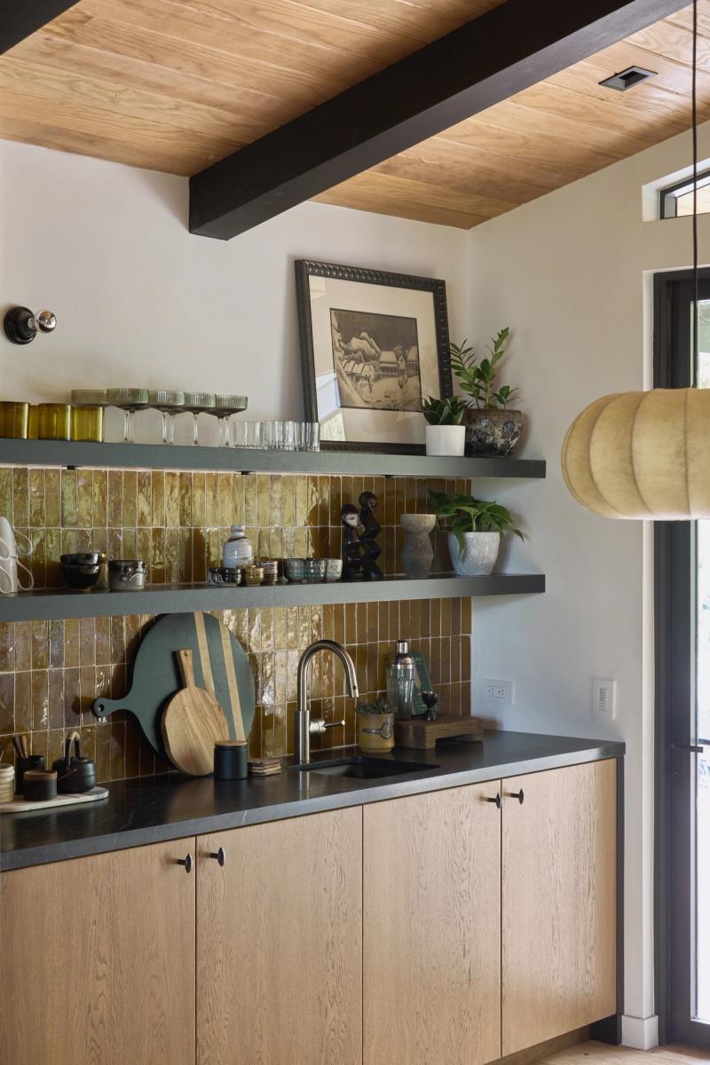 A stylish wet bar area featuring light oak cabinetry with dark stone countertops, iridescent amber zellige tile backsplash, floating dark shelves styled with glassware and plants, and a wood-paneled ceiling with black beams.