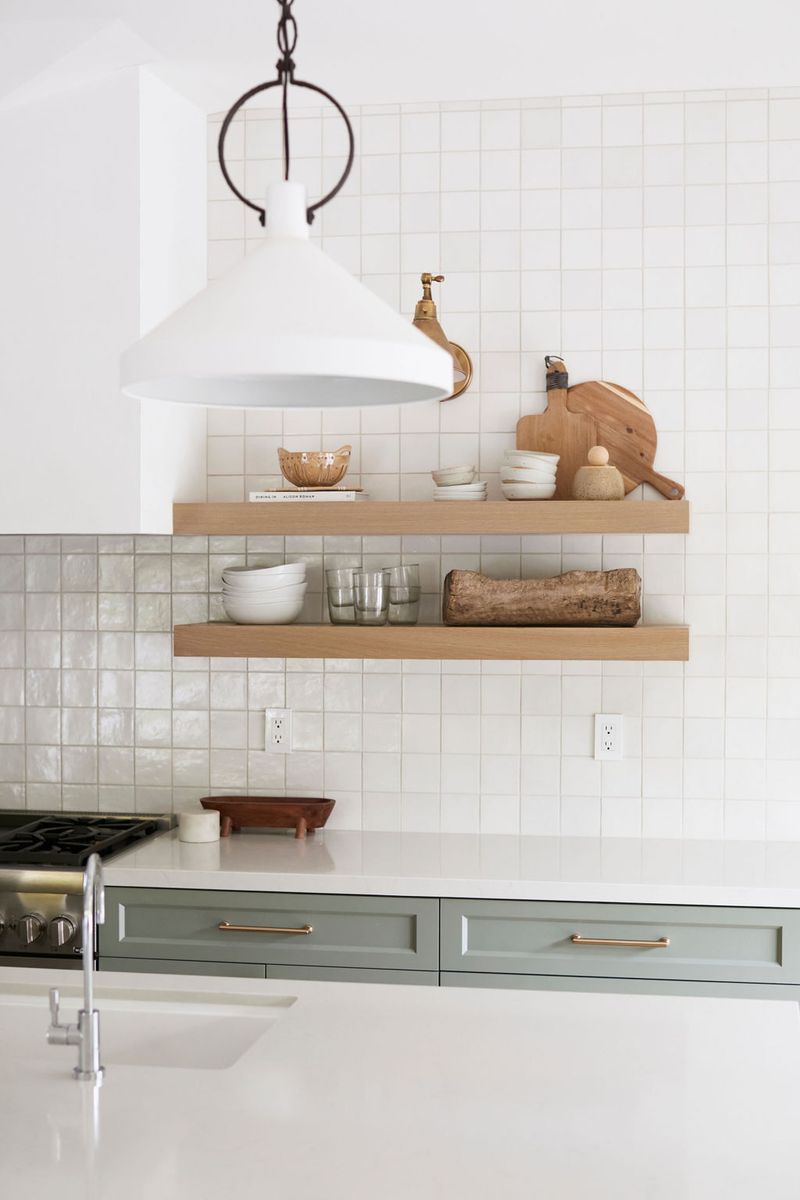 A bright, modern kitchen featuring sage green lower cabinets with brass hardware, two floating oak shelves styled with ceramics and cutting boards, white square tile backsplash, and a white pendant light.