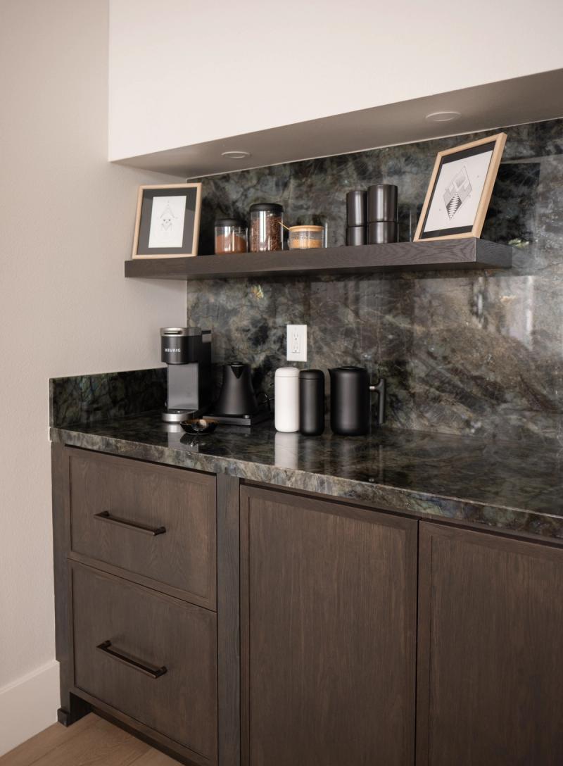 A built-in coffee bar featuring dark stained wood cabinetry with bar pulls, a dramatic green labradorite stone countertop and full-height backsplash, open floating shelf with decorative jars and framed art, and a Keurig coffee maker on the counter.