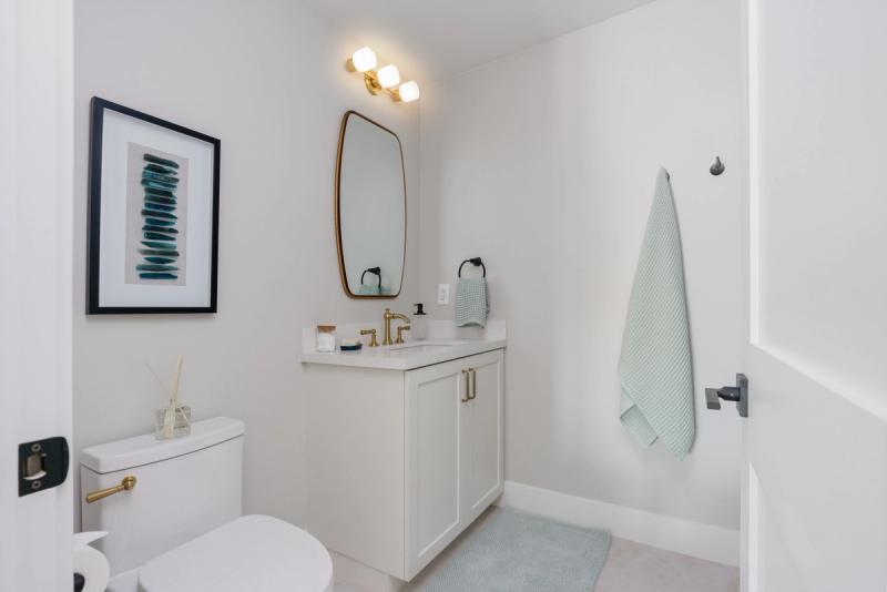 A bright, modern powder room featuring a white single-sink vanity with brass fixtures and cabinet pulls, an oval gold-framed mirror, and coordinating mint green towels and rug.