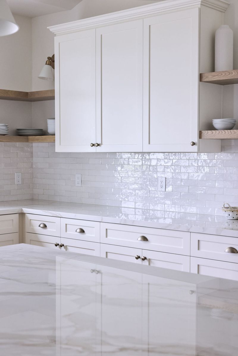 A bright, all-white kitchen featuring shaker-style cabinetry with brushed nickel hardware, marble countertops, a glossy white zellige tile backsplash, and floating oak shelves on either side of the upper cabinet.