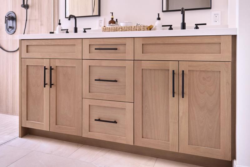 A double bathroom vanity built from light natural wood with shaker-style cabinet doors and drawers, black bar pulls, white countertop, and dual black faucets with undermount sinks.