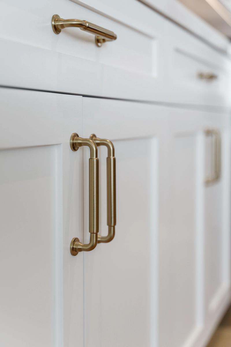 A close-up of white shaker-style kitchen cabinets featuring elegant double-bar brushed brass pulls on the cabinet doors and a matching brass pull on the upper drawer.