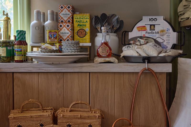 A close-up of a light oak kitchen sideboard or counter displaying an assortment of gourmet pantry items including a paella kit, Maldon salt, olive oil, and woven baskets stored below.