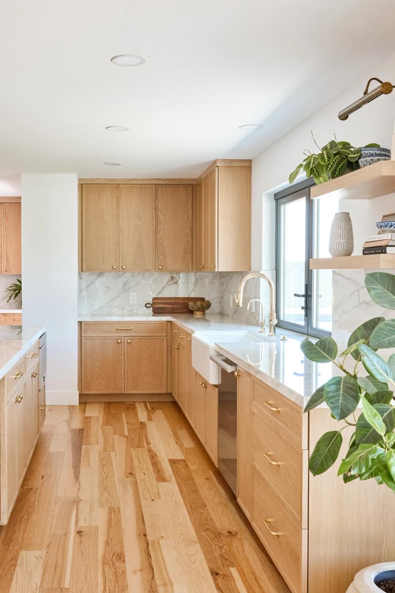 A bright, modern kitchen featuring custom white oak shaker cabinets with brass hardware, marble countertops and backsplash, a farmhouse sink, and light hardwood floors. Open shelving near a window holds decorative plants and books.