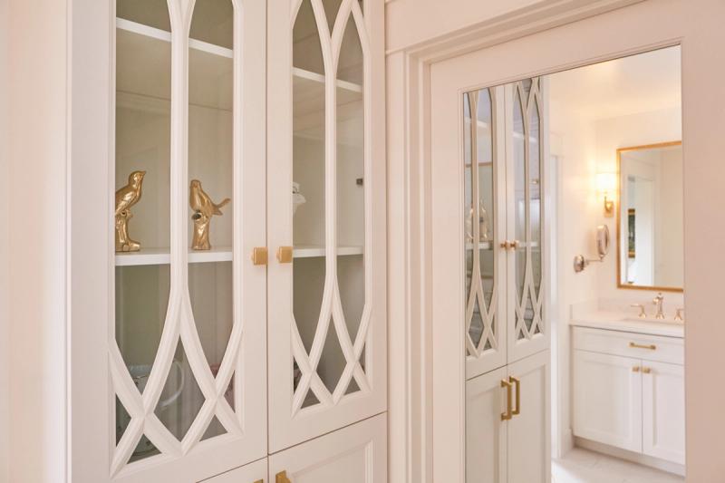 A floor-to-ceiling white built-in cabinet with decorative arched glass door panels and brass hardware, displayed alongside a glimpse into a bright white bathroom with gold-framed mirror and vanity.