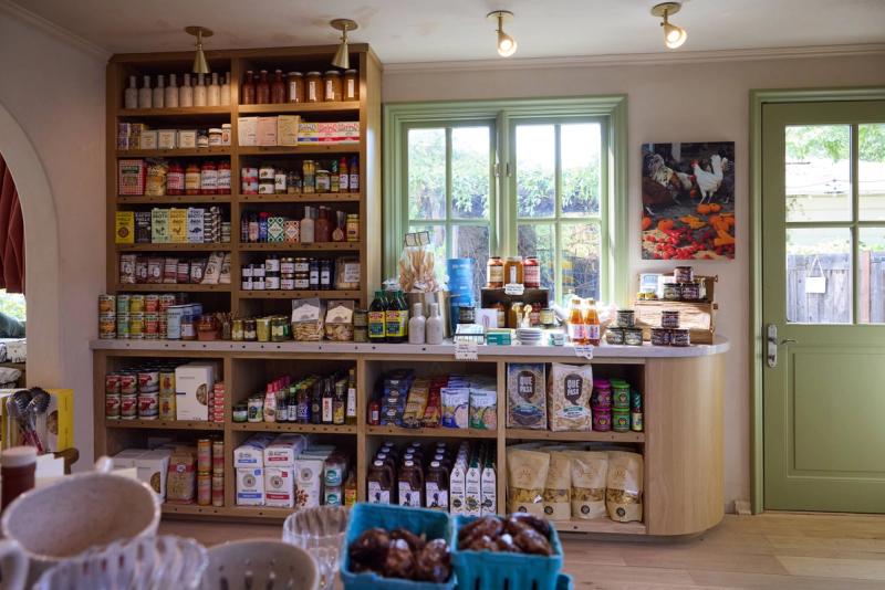 A warmly lit specialty food shop interior featuring custom oak built-in shelving stocked with pantry goods and a curved display counter, with green-trimmed windows overlooking a garden.