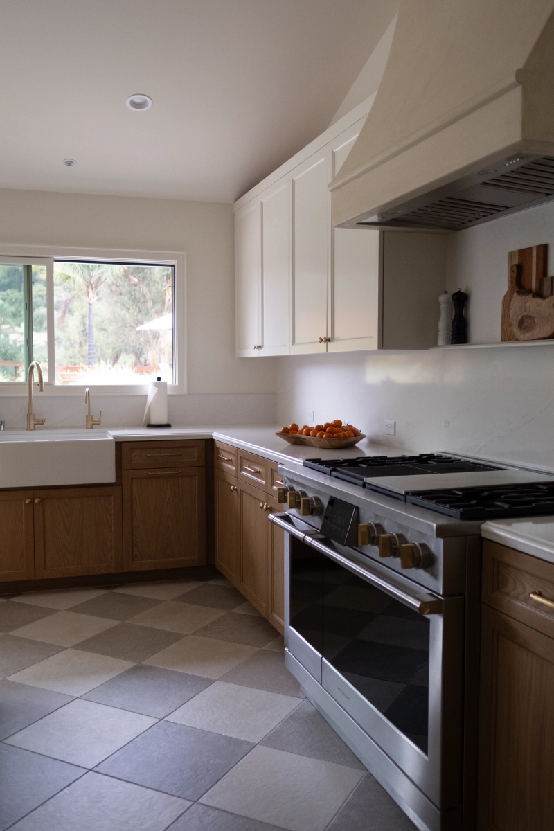 A bright, modern kitchen featuring warm oak lower cabinets and white upper cabinets, a farmhouse sink, stainless steel professional gas range with brass hardware, white quartz countertops, and a checkered tile floor.