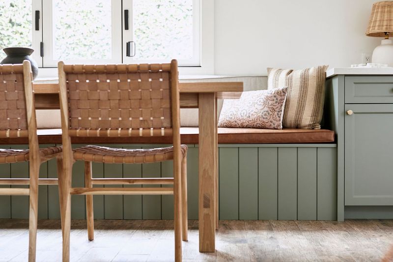 A cozy dining nook featuring a sage green built-in storage bench with a leather cushion and decorative pillows, paired with woven leather and wood dining chairs around a light wood table.