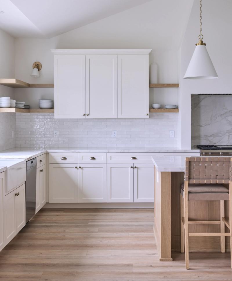 A bright, airy kitchen featuring cream shaker-style cabinetry, white subway tile backsplash, light oak open shelving, brass hardware and lighting, white quartz countertops, and light wood flooring with a woven leather bar stool at the island.