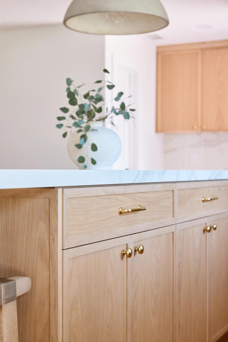 A close-up of a light white oak kitchen island featuring brass pulls and knobs, topped with a white marble countertop. A pale blue vase with eucalyptus branches sits on the counter in the background.