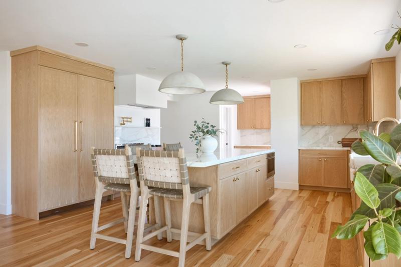 A bright, modern kitchen featuring light oak cabinetry, a large center island with white marble countertop, woven leather bar stools, sage green pendant lights, and hardwood floors.