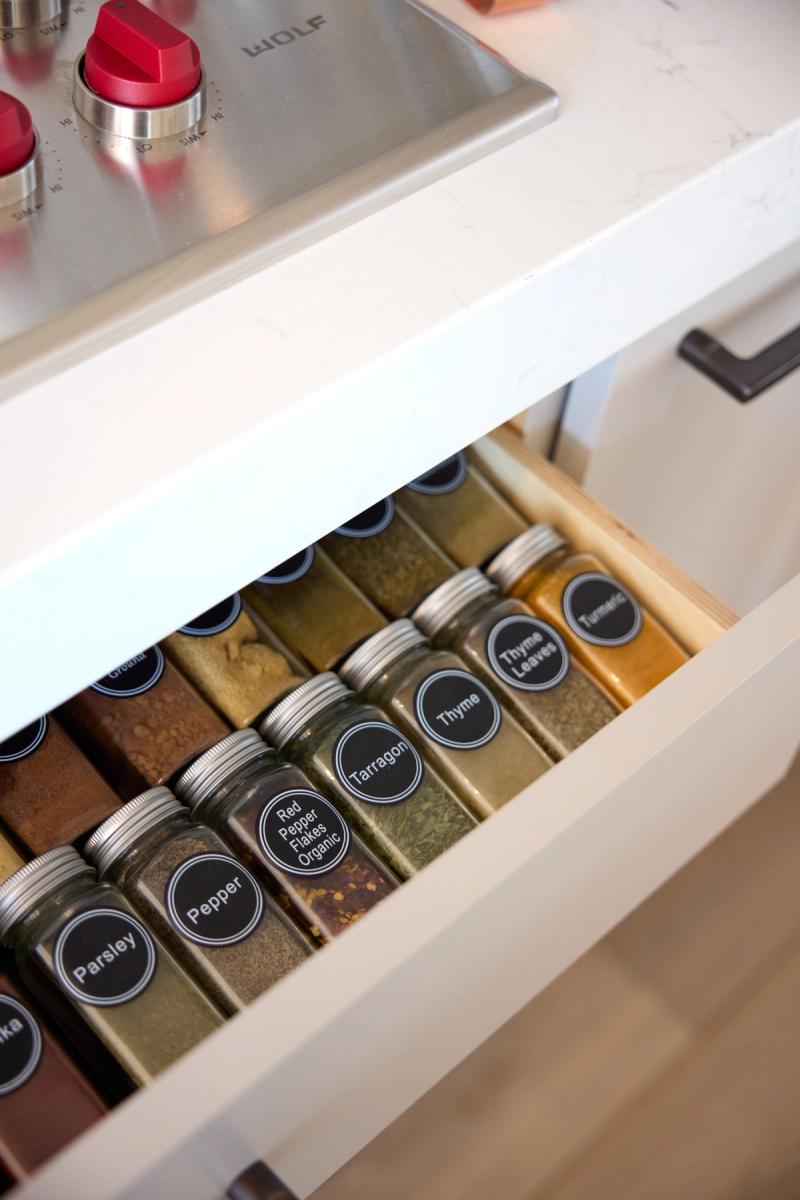 An open kitchen drawer beneath a Wolf range cooktop reveals neatly organized rows of labeled glass spice jars with black chalkboard-style lids, set in a white cabinetry unit with a marble countertop.