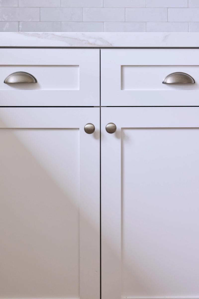 Close-up of white shaker-style bathroom vanity cabinets with brushed nickel cup pulls on the upper drawers and round knobs on the lower cabinet doors, topped with white marble countertop and subway tile backsplash.